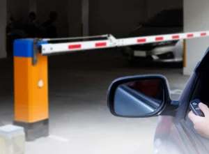 Automatic barriers at a car park entrance with a raised barrier arm and driver using a remote control.