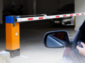 Automatic barriers at a car park entrance with a raised barrier arm and driver using a remote control.