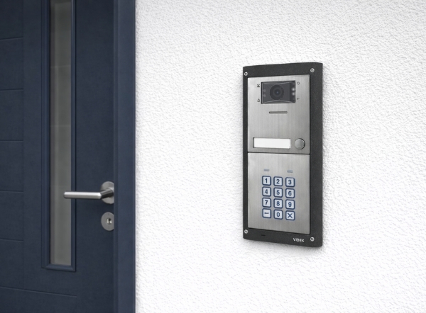 Video intercom mounted on a white exterior wall beside a modern front door, featuring a camera and keypad, ideal intercoms for home security and access control.