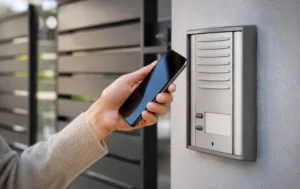 Hand holding a smartphone near a modern access control gsm intercom mounted beside a building entrance.